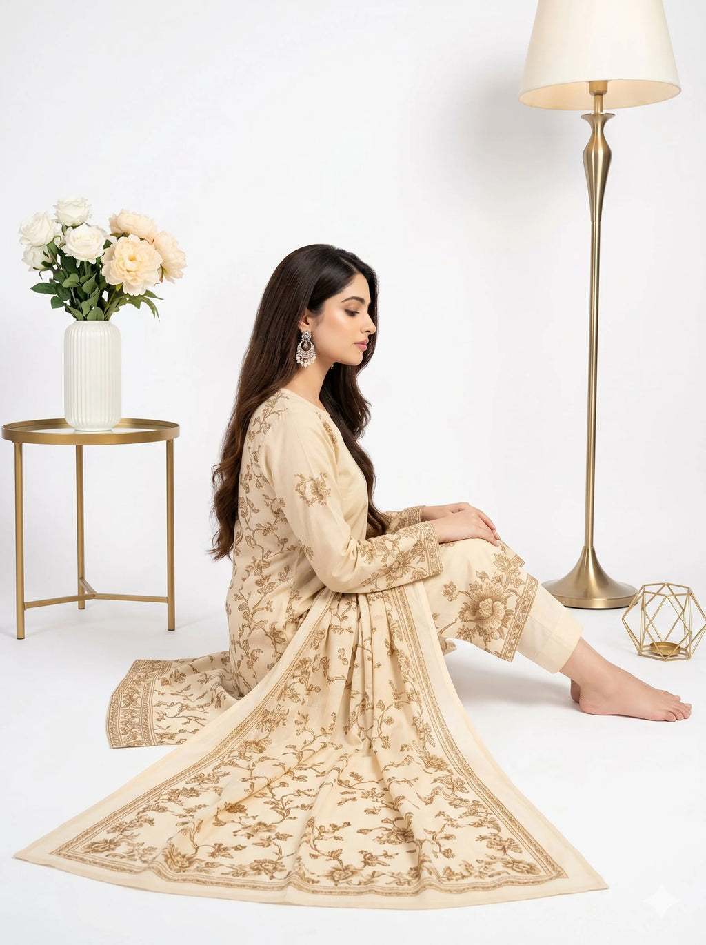 Woman in a beige embroidered outfit sitting on the floor with a white background